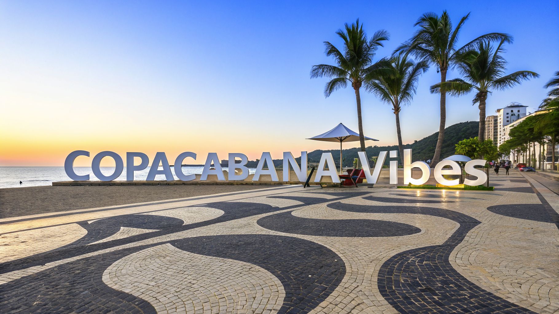 Large 'COPACABANA Vibes' sign on the mosaic promenade of Copacabana beach at sunset, with palm trees and ocean.